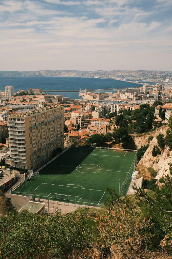 Vue panoramique de Marseille avec un terrain de football urbain, les immeubles du centre-ville et la mer Méditerranée en arrière-plan sous un ciel bleu