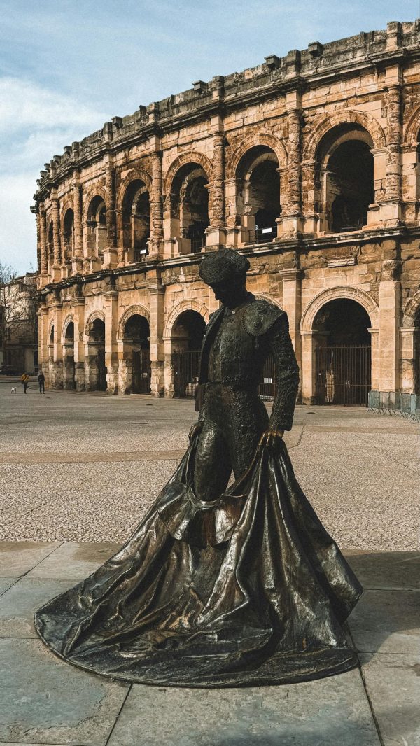 Statue de torero en bronze devant les Arènes de Nîmes, amphithéâtre romain emblématique sous un ciel dégagé